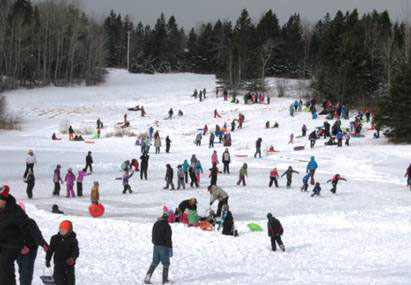 Many families ce skating  on a pond with others sliding on the snowy hill beyond it.