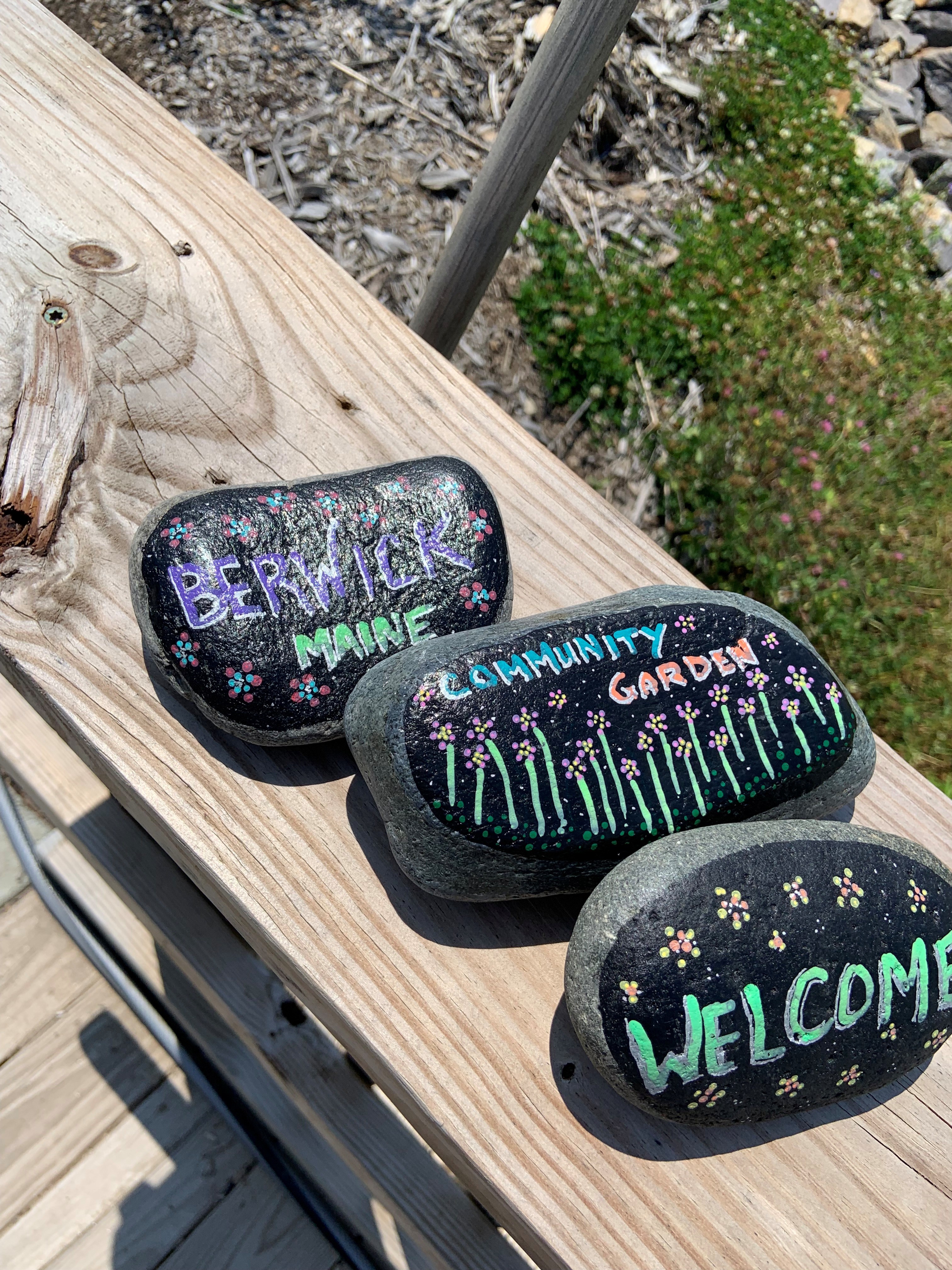 Painted stones reading "Berwick Maine," "Community Garden," and "Welcome" on a wooden plank.